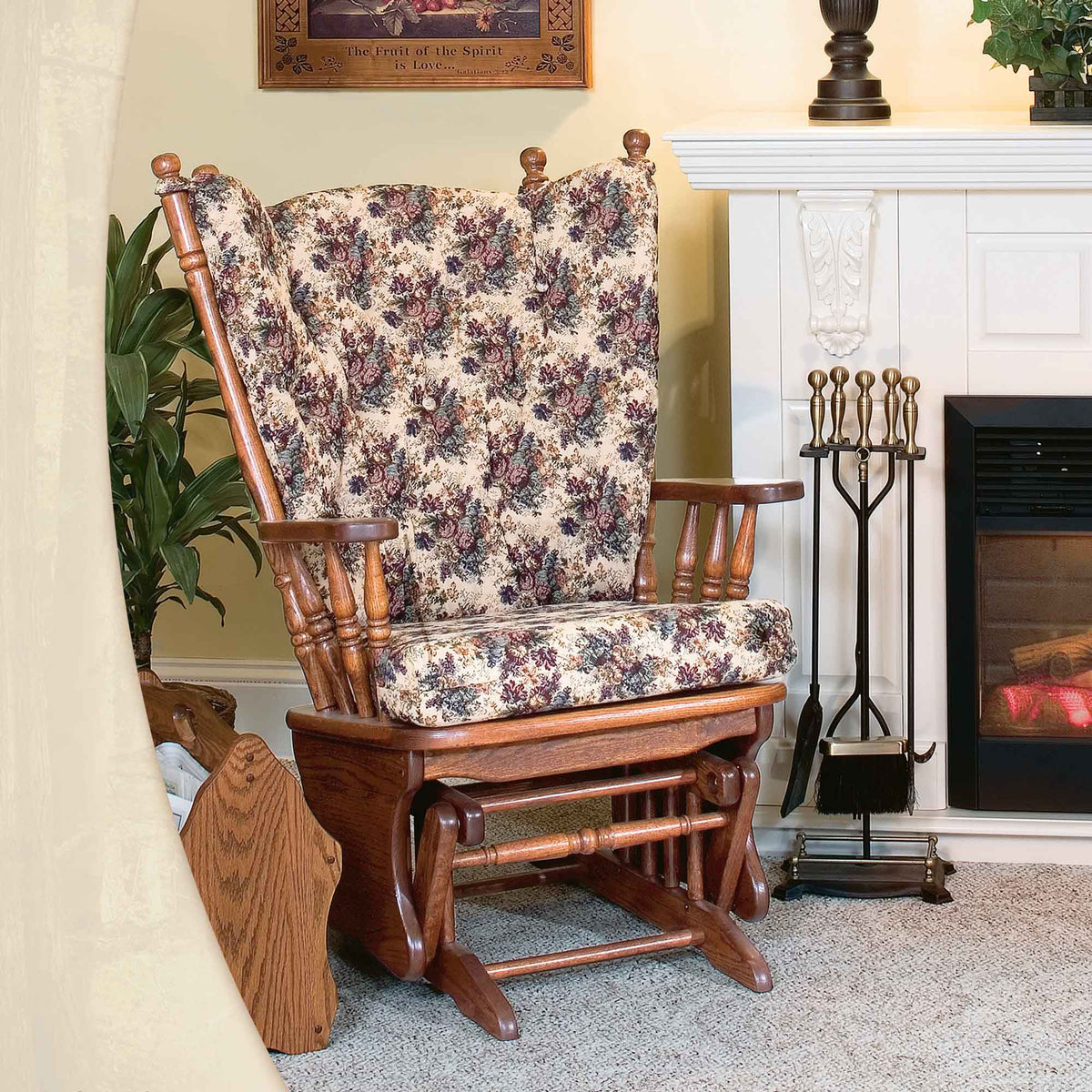 Solid oak glider rocker with wing-style floral tufted back cushion photographed by a fireplace with brass tool set