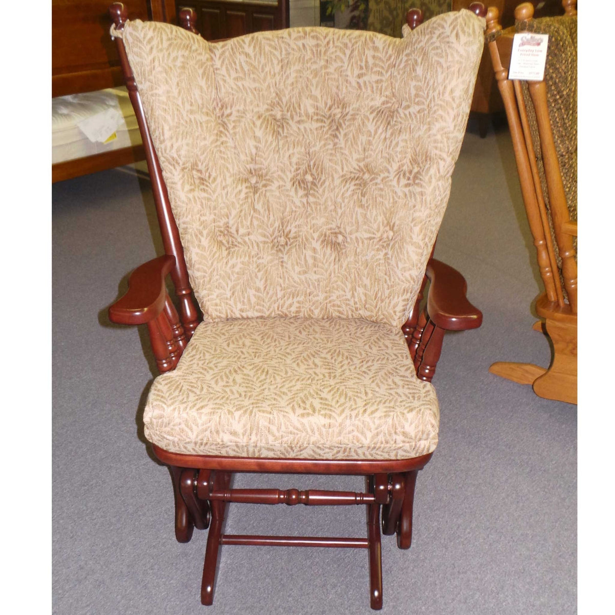 Front view of the cherry-finished glider rocker with tan leaf-pattern tufted cushions in the furniture showroom