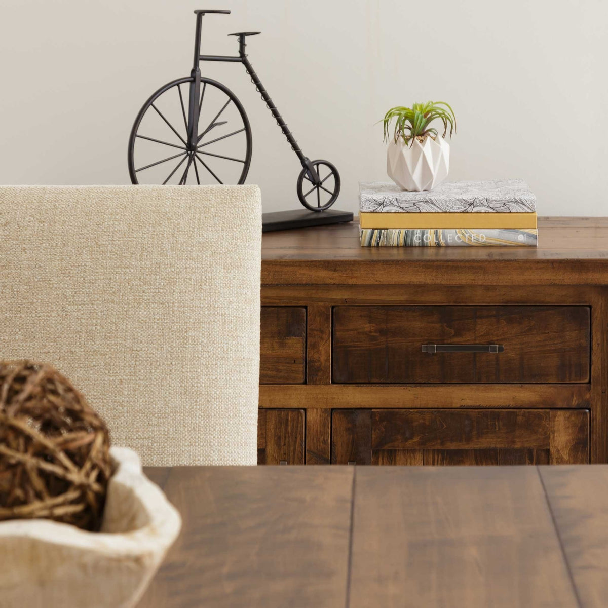 Corner of a rustic plank dining table with a linen upholstered chair back and a wood sideboard behind