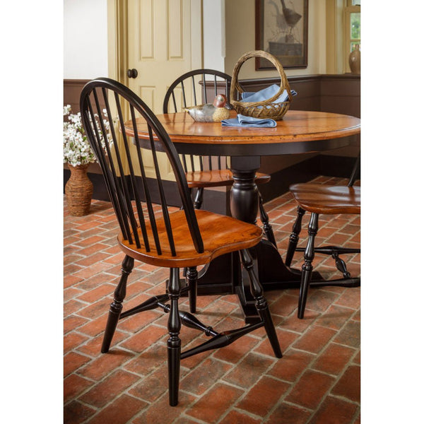 Round pedestal dining table with a cherry top and black base surrounded by bow back Windsor chairs on a brick floor