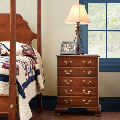 Five-drawer cherry bedside chest with brass bail pulls and ogee bracket feet beside a poster bed and lamp