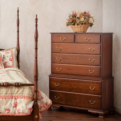 Cherry chest on chest with seven drawers and brass bail pulls beside a turned-post bed, basket of flowers on top