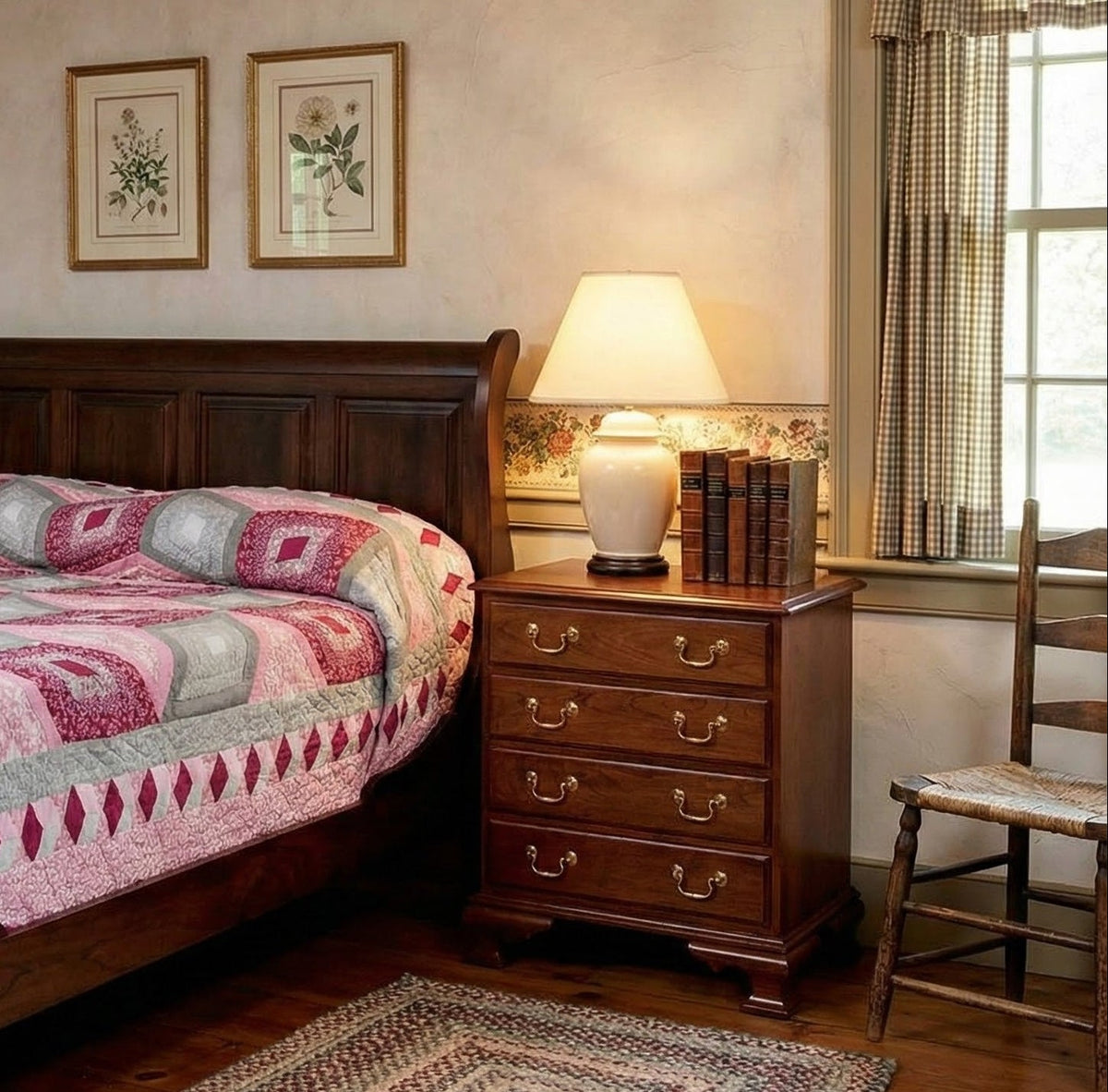 Cherry sleigh bed headboard beside a four-drawer nightstand with brass pulls, lamp and books by a checked curtain