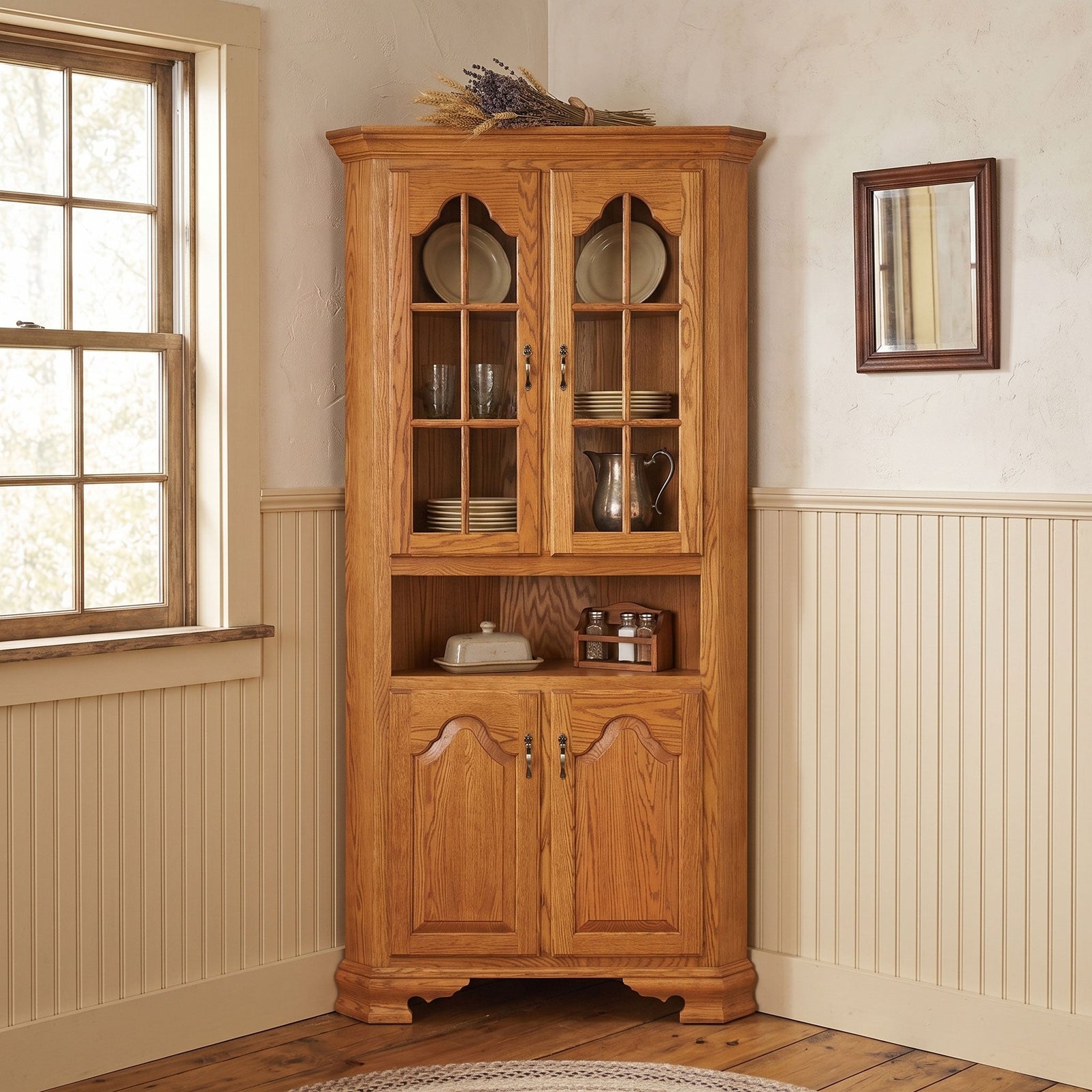 Oak corner china cabinet with arched mullioned glass doors filled with dishes, open center shelf and raised panel doors below