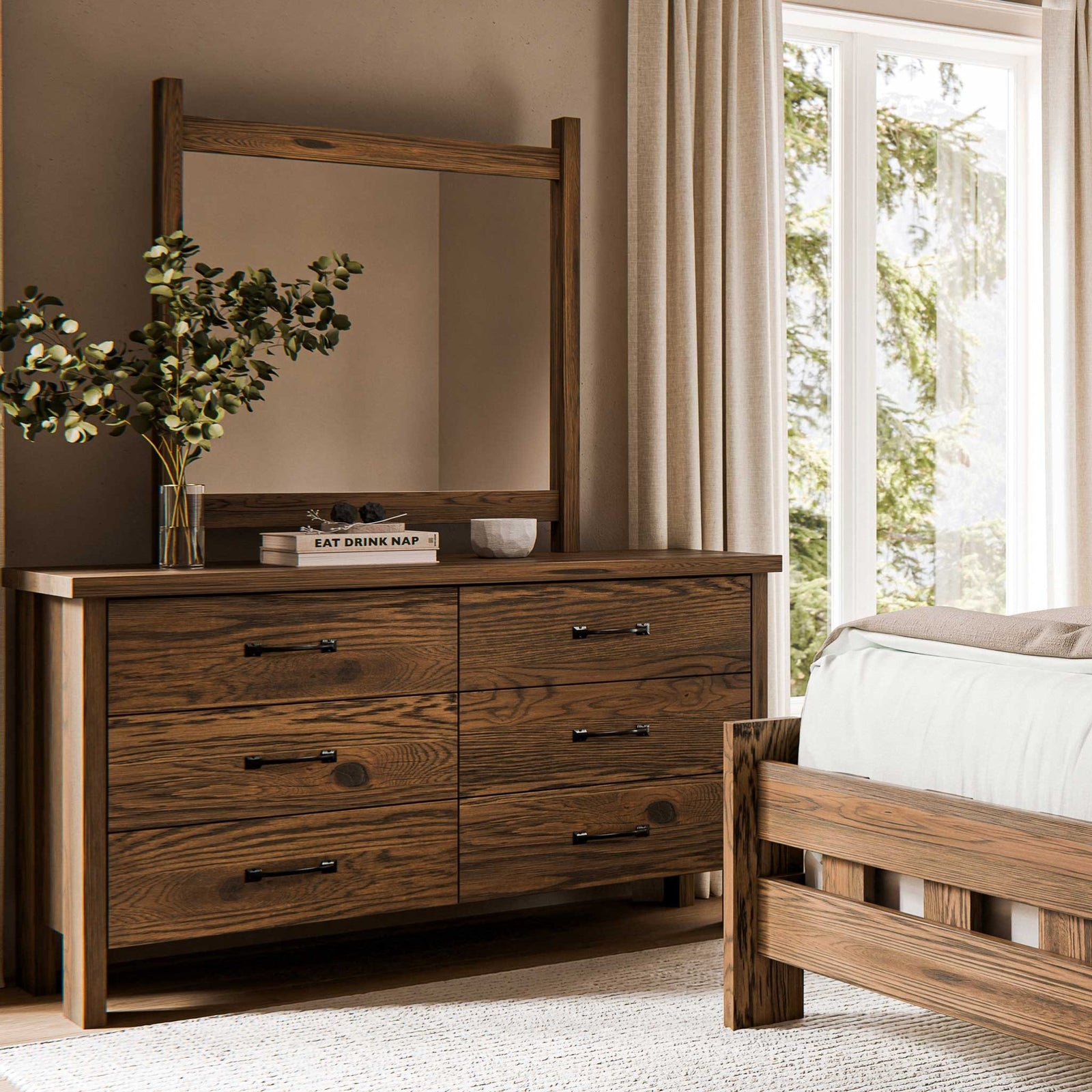 Oak six drawer dresser with dark pulls topped with a mirror, books and greenery in a warm bedroom