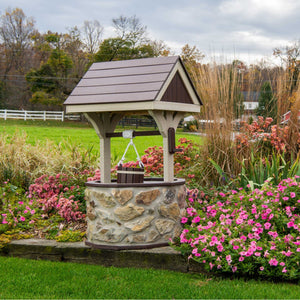 Stone-base wishing well with brown poly roof and hanging bucket, set in a landscaped bed of pink petunias and sedum