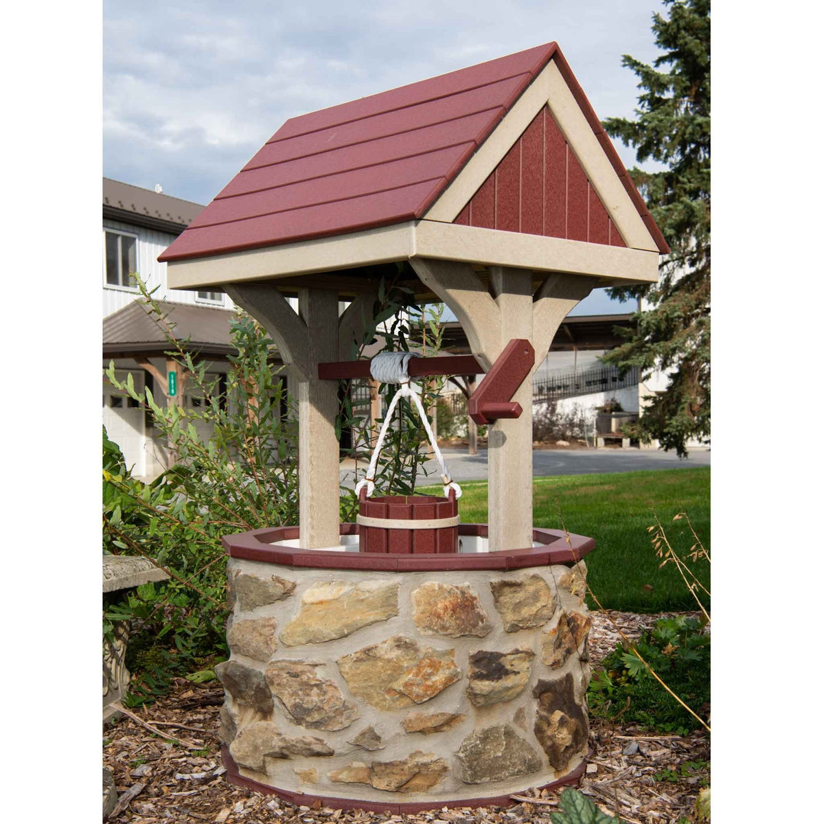 Front view of a stone wishing well with red gable roof, tan trim, crank handle and red rope bucket in a mulched bed