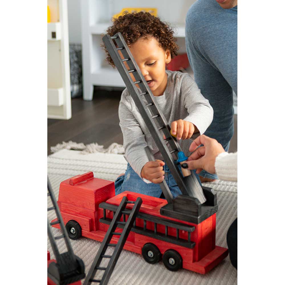 Toddler and adult playing on a rug with a red wooden fire truck toy, raising its long black extension ladder