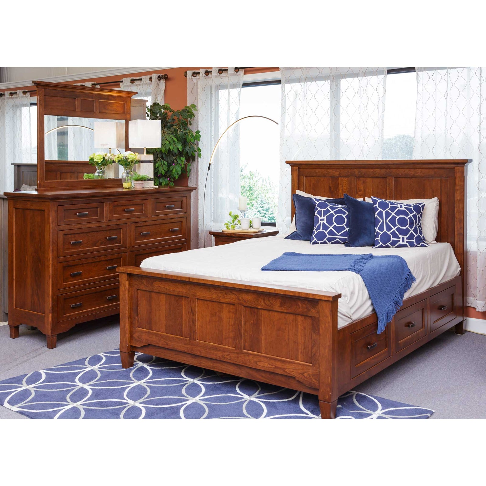 Cherry bedroom set styled in a showroom with a panel storage bed, blue bedding, nine-drawer dresser and mirror