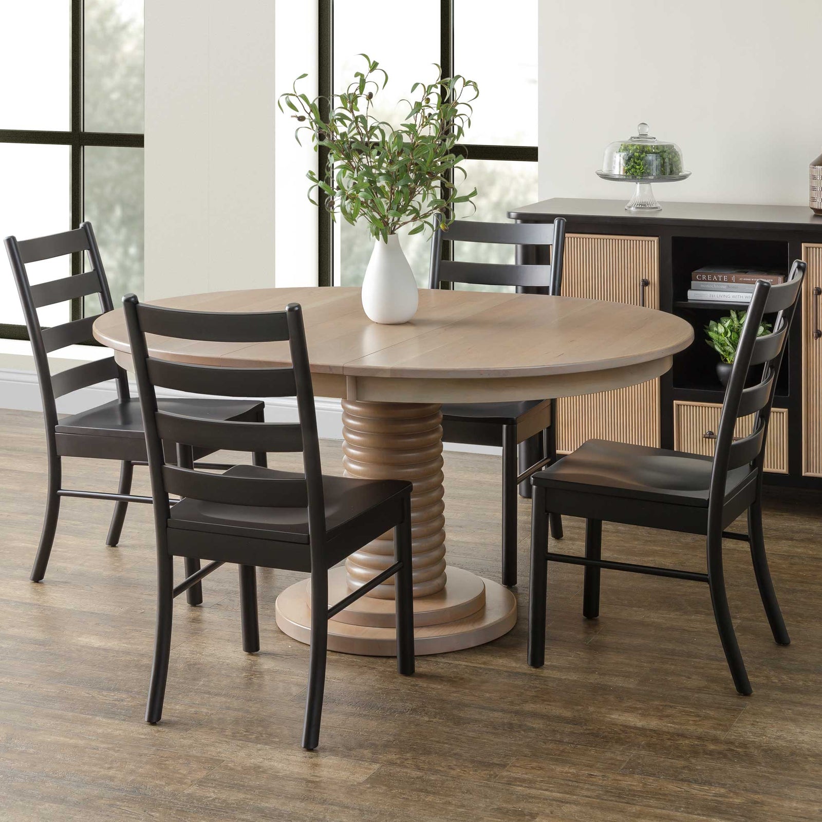 Round light oak pedestal dining table with a ribbed turned column base and four black ladder-back chairs