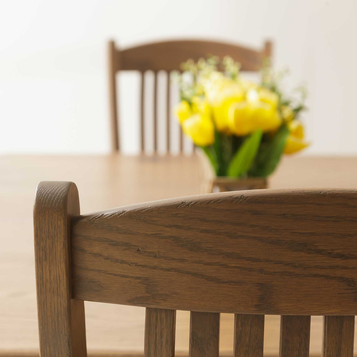 Close-up of the chair's curved crest rail and back slats showing open oak grain, tulips blurred behind