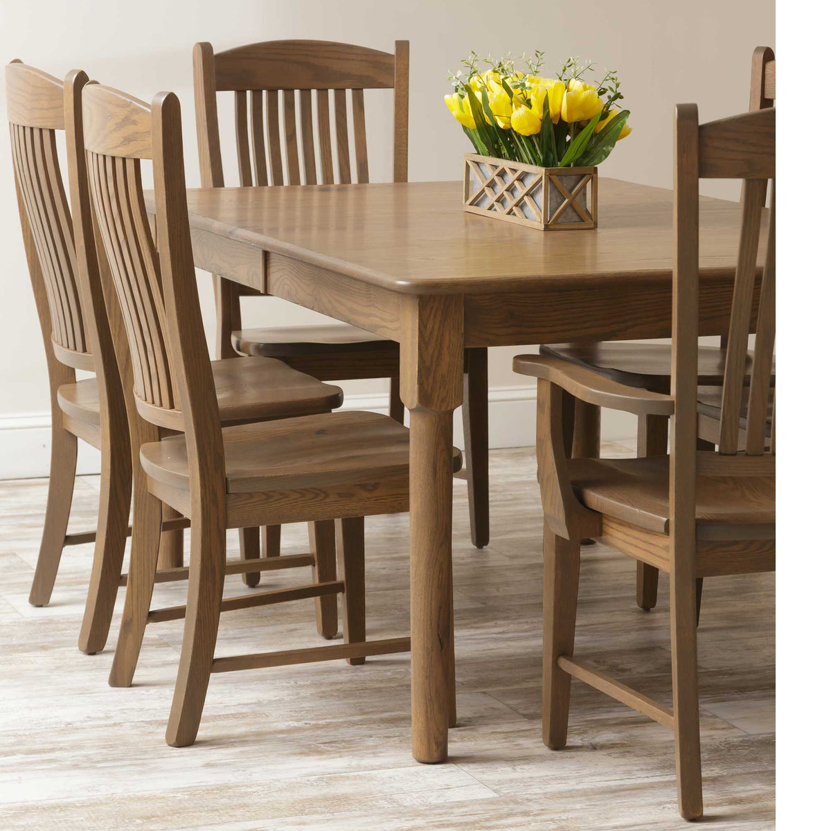 Rectangular oak dining table surrounded by six slat-back chairs with yellow tulips as a centerpiece