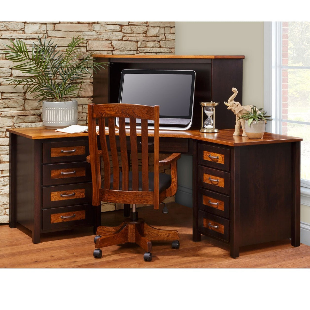 Two-tone corner L desk with black painted base, oak tops and drawer panels, hutch top with monitor and matching swivel office chair in a stone-walled office