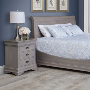 Gray finished three-drawer nightstand with dark cup pulls and bracket feet beside a sleigh bed in a blue bedroom