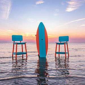 Sunset shot of a surfboard shaped bar table standing on end between two aqua bar stools in the water