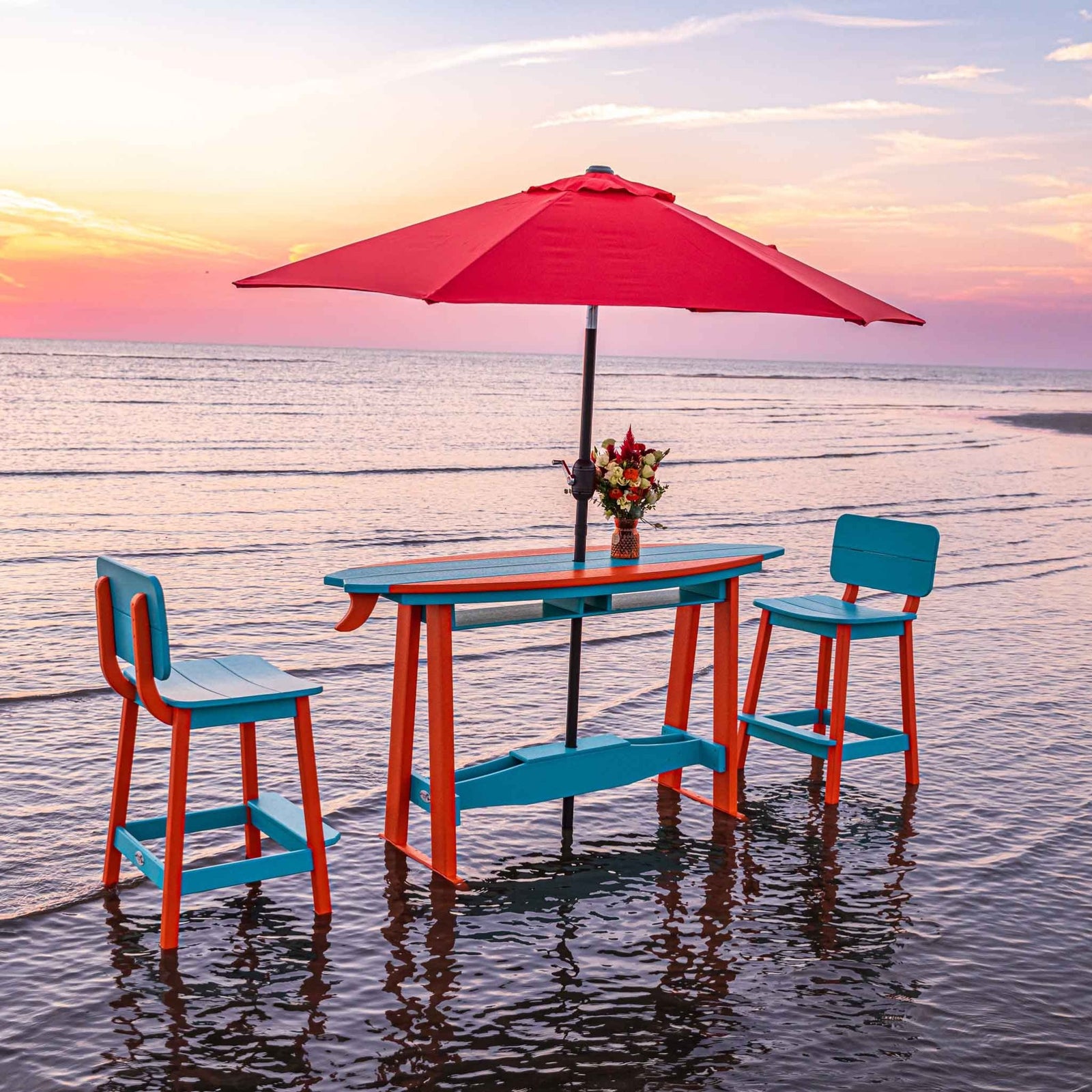 Aqua and orange surfboard bar table with two bar stools and a red umbrella standing in shallow surf
