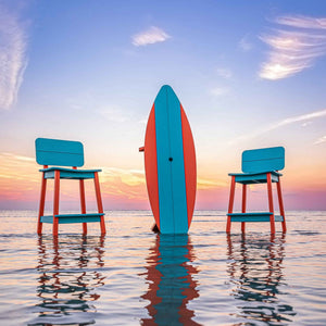 Upright orange and turquoise surfboard table between two matching bar stools in calm sea water