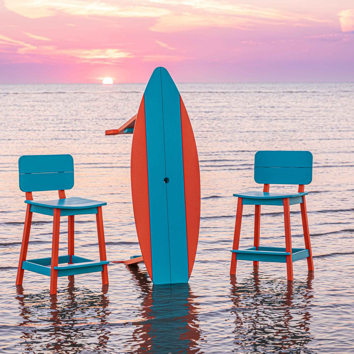 Surfboard bar table stood upright between two turquoise bar stools in the ocean at sunset