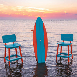 Two turquoise and orange poly bar stools flanking an upright surfboard table in the sunset surf