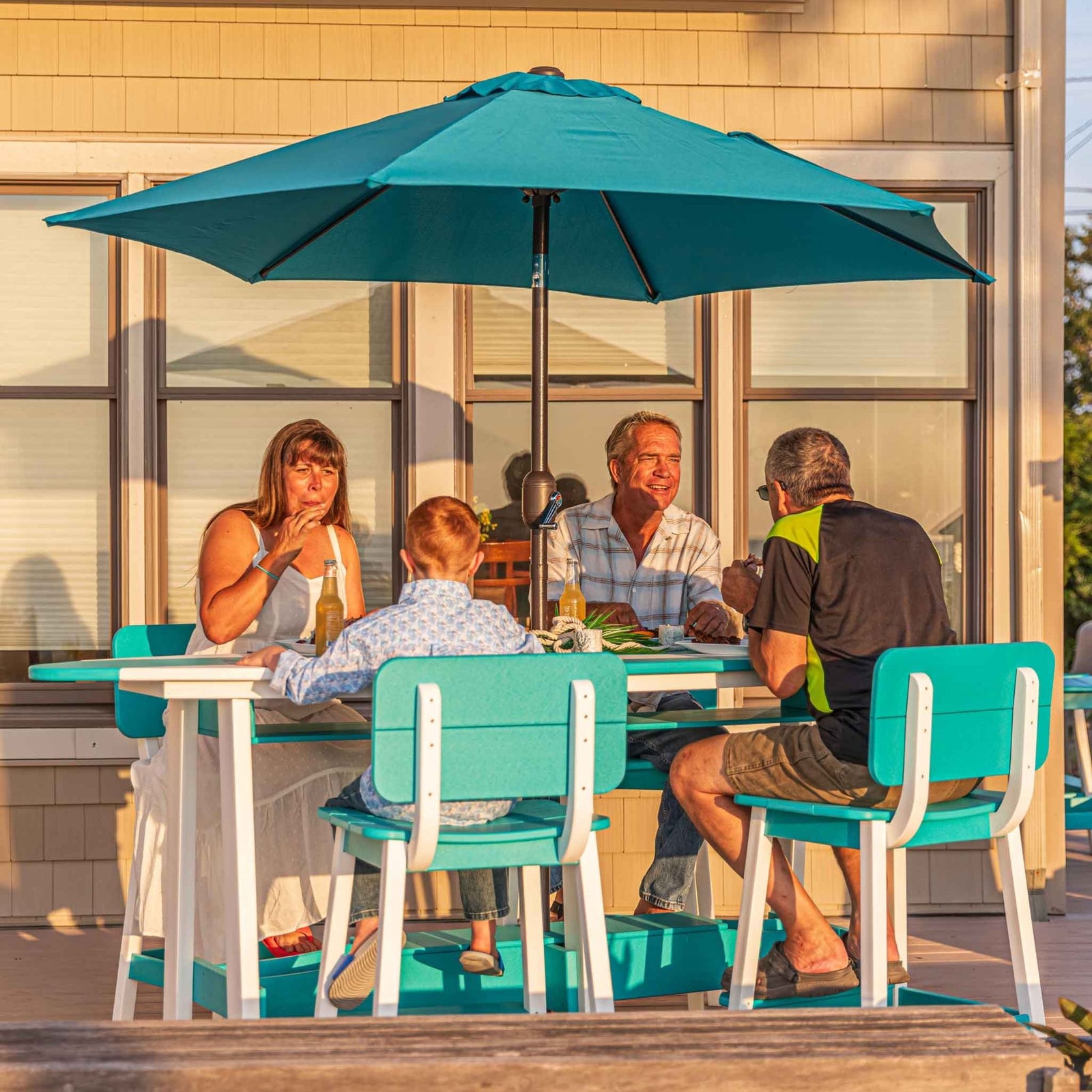 Family dining at a turquoise and white surfboard shaped bar table under a teal umbrella on a beach house deck