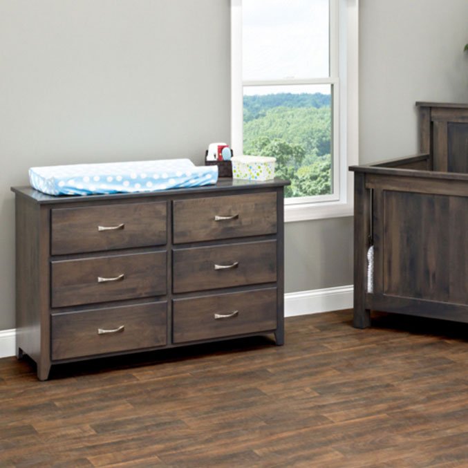 Gray six-drawer dresser topped with a blue changing pad beside a matching crib in a nursery with a bright window