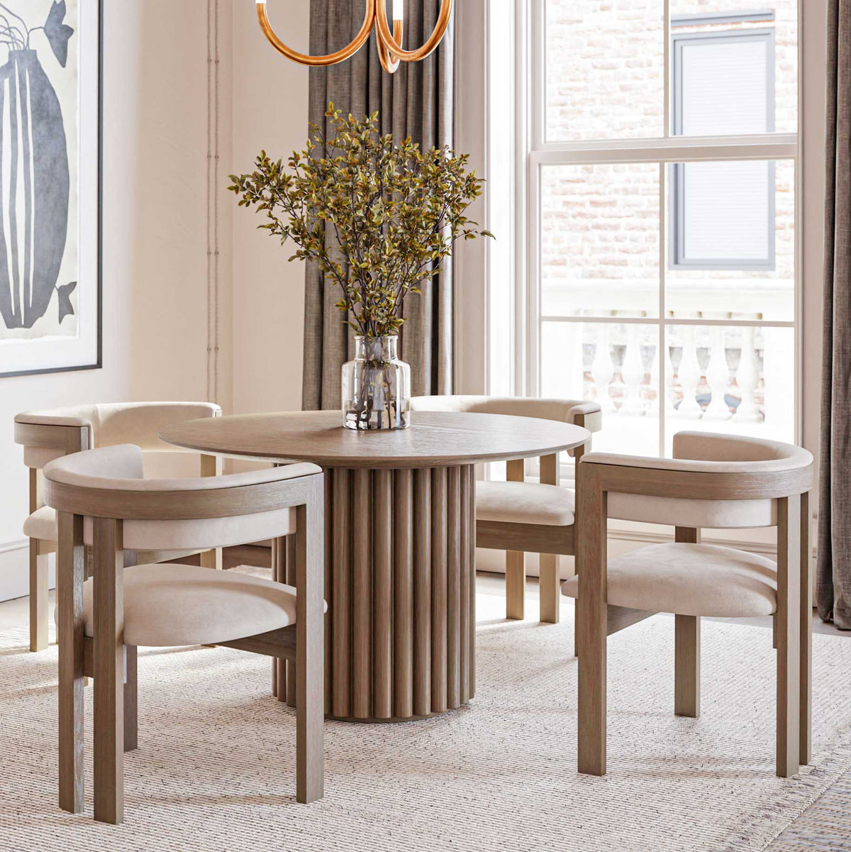 Round fluted pedestal dining table in gray washed oak with four cream barrel back arm chairs in a sunny dining room