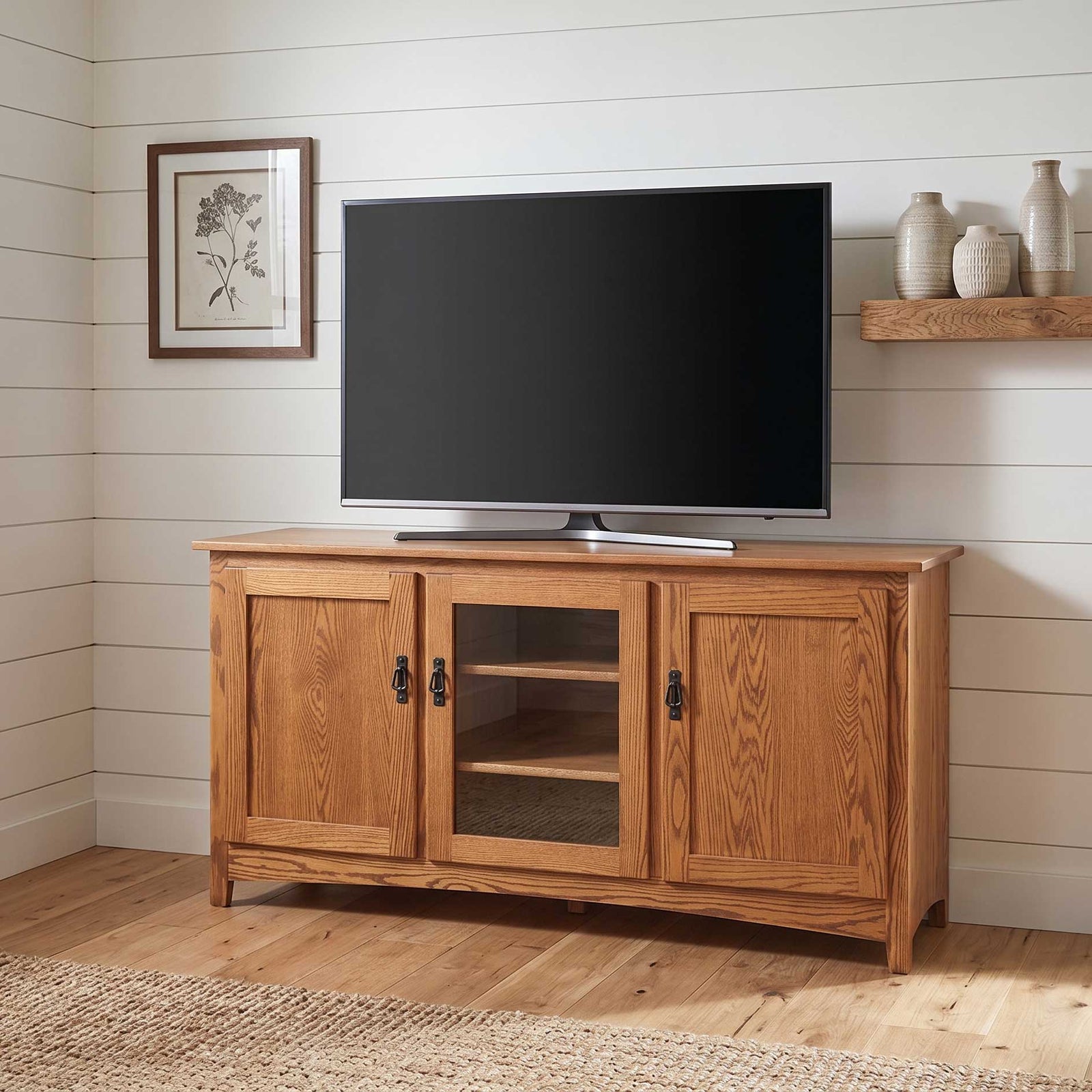 A rustic medium-brown wooden TV stand cabinet with two doors, one glass-paneled door, and open shelving, shown holding a flat-screen television in a shiplap-walled room.