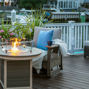 Gray poly swivel glider with tan cushions and blue pillow beside a lit fire pit table on a marina deck at dusk