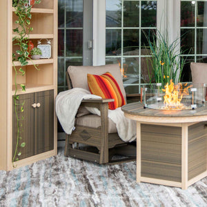 Driftwood gray poly swivel glider chair with tan cushions and a throw beside a lit round gas fire pit table