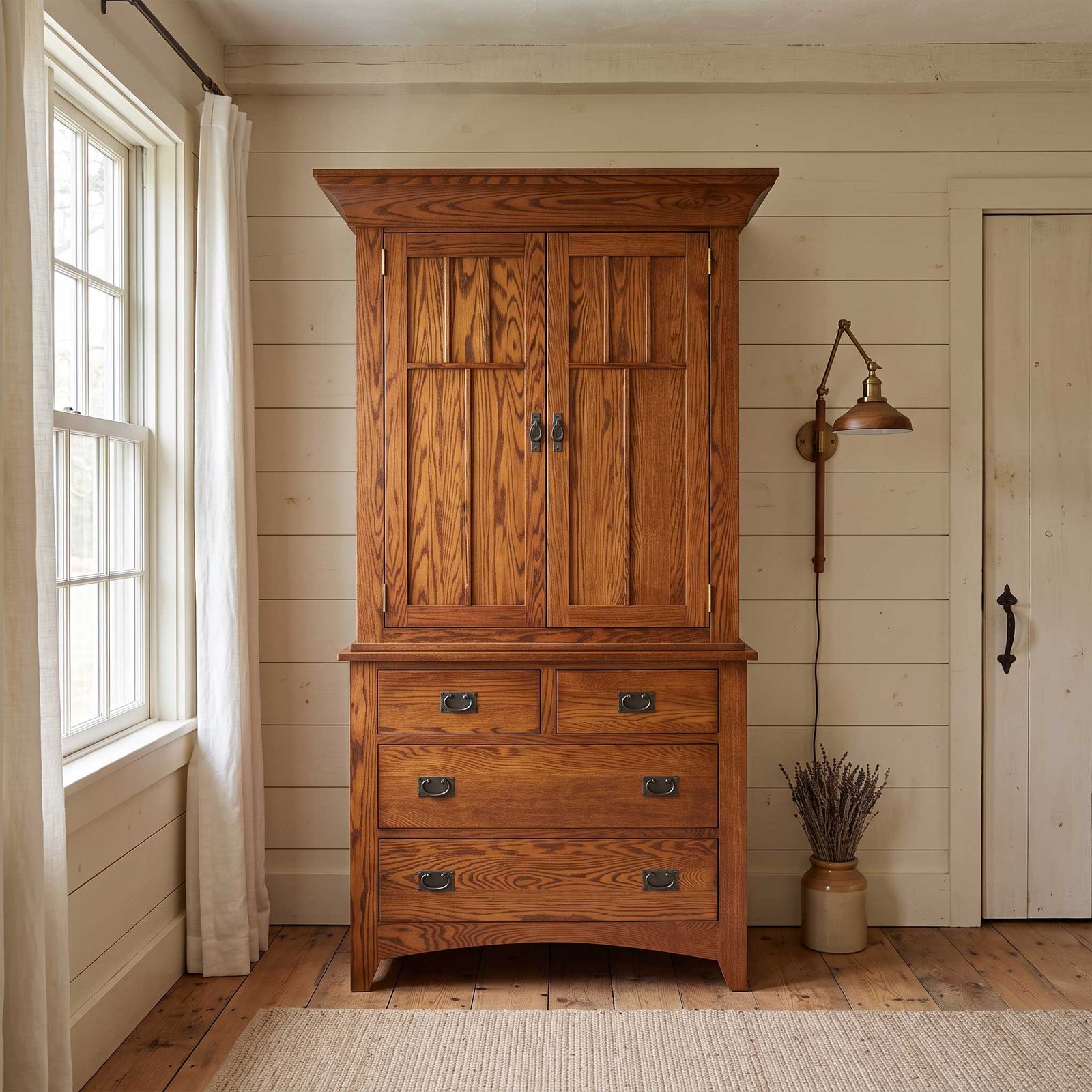 A rustic wooden armoire cabinet with a two-door upper section and three drawers below, styled in a bright, neutral room with a window, wall lamp, and door.