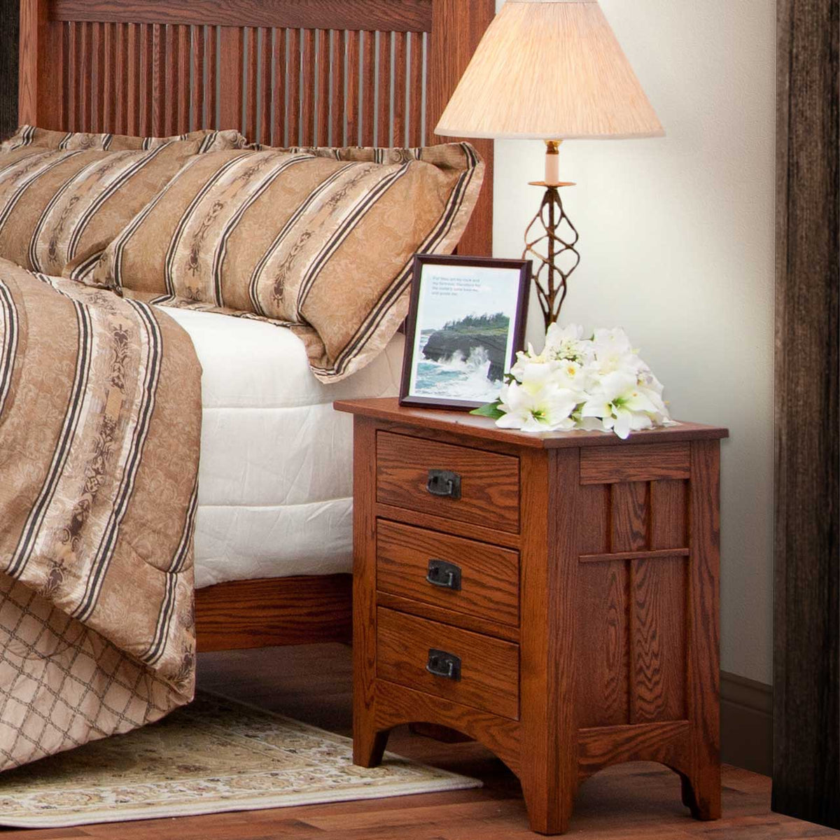 Oak Mission three-drawer nightstand with slatted side panel and black bail pulls beside a slatted bed and lamp
