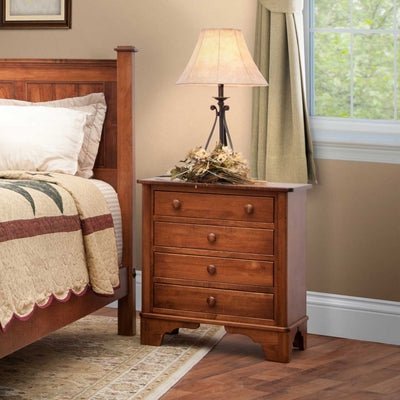 Four-drawer cherry nightstand with round wood knobs beside a paneled bed, holding a lamp and dried flowers