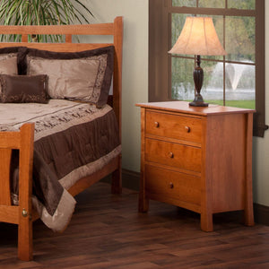 Cherry three-drawer bedside chest with round knobs and flared legs beside a bed, holding a table lamp