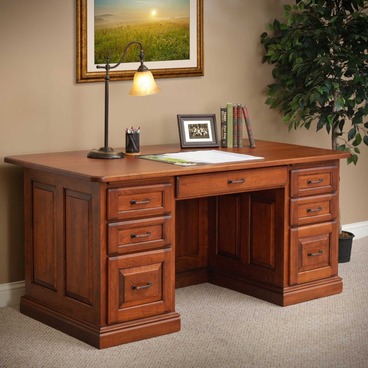 Cherry executive desk with lamp and books, framed sunrise painting above and a ficus tree beside it