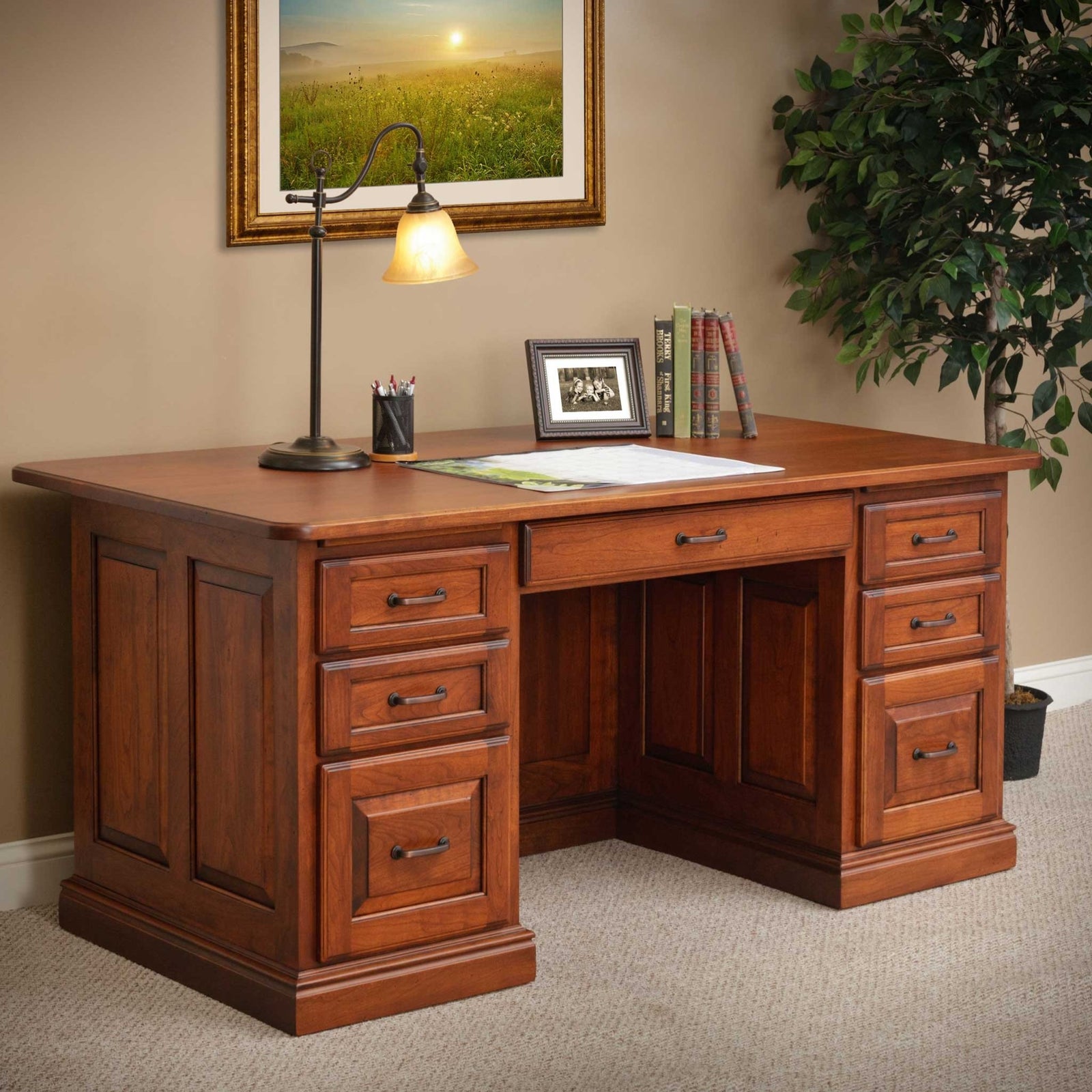 Cherry executive desk with lamp and books, framed sunrise painting above and a ficus tree beside it
