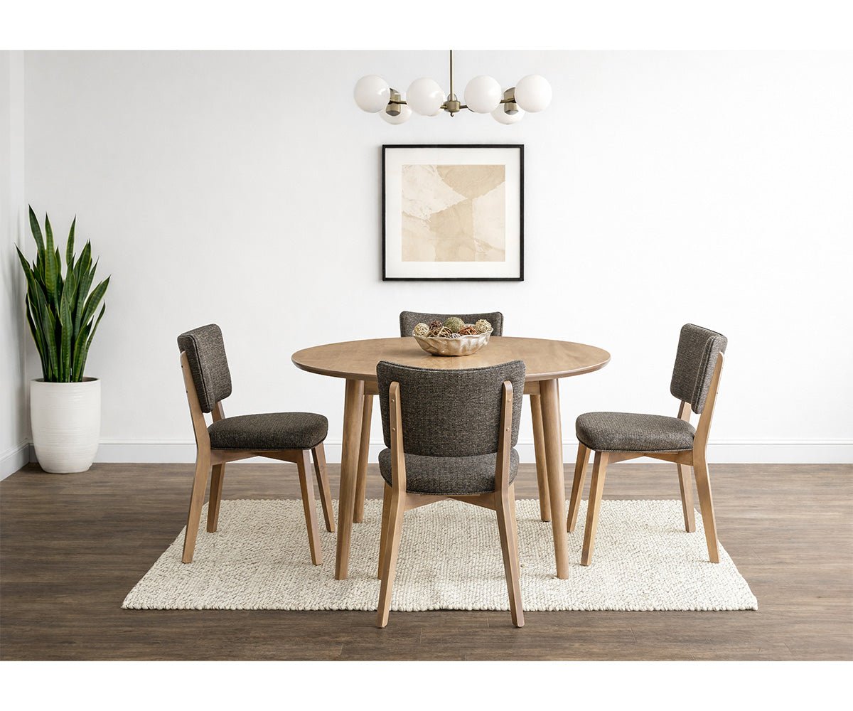 Round oak dining table with four gray cushioned chairs on a woven rug under a brass globe light fixture