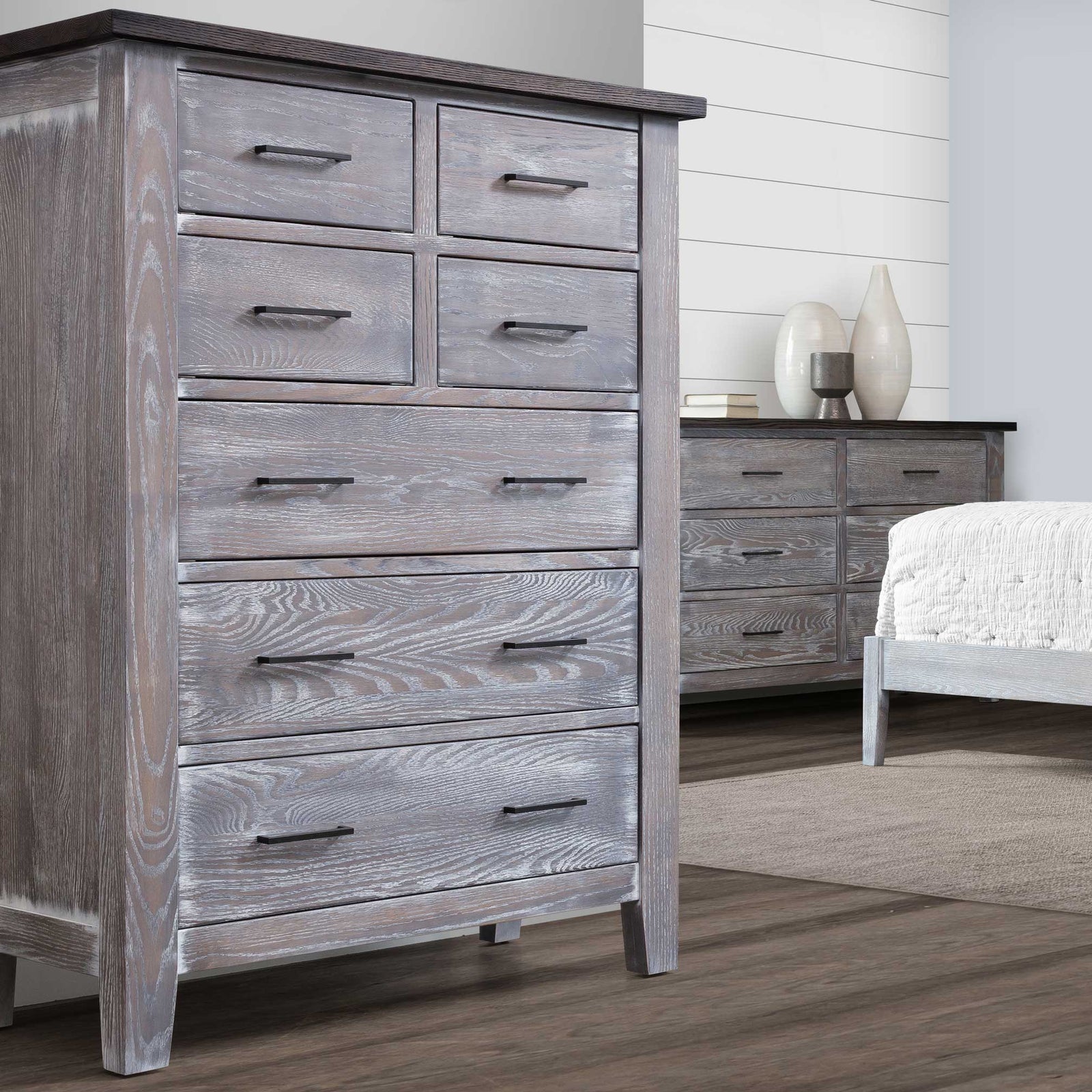 Tall gray oak chest of drawers with seven drawers and black bar pulls, staged in a bedroom next to a matching dresser