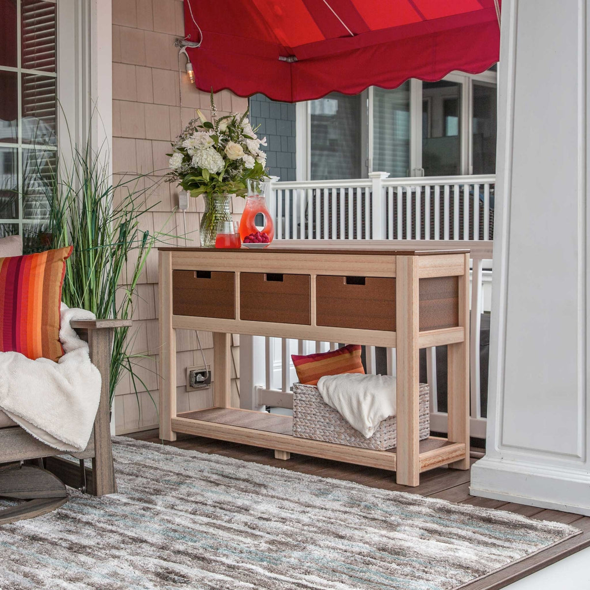 Light poly console table with three drawers and a lower shelf holding a basket, styled with flowers on a porch