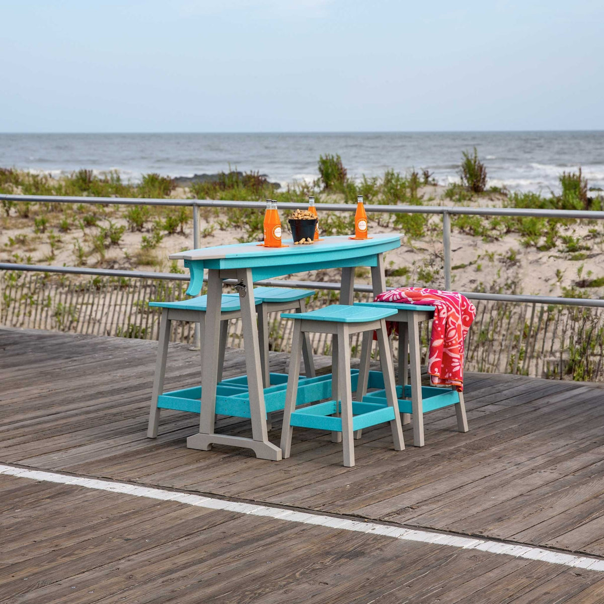 Turquoise poly surfboard shaped counter height table with gray legs and stools on an oceanfront boardwalk