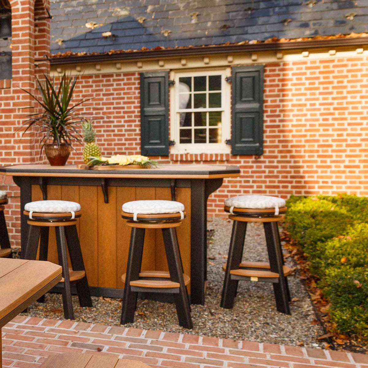 Three cedar and black poly swivel stools with round cushion pads at an outdoor bar by a brick house