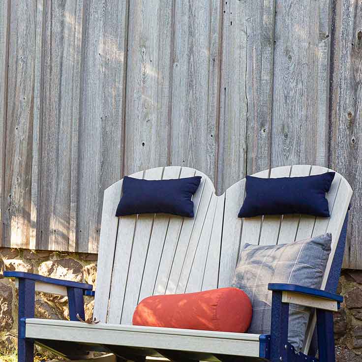 Navy neck pillows and a rust cylinder bolster on a weatherwood Adirondack loveseat by a barn wall