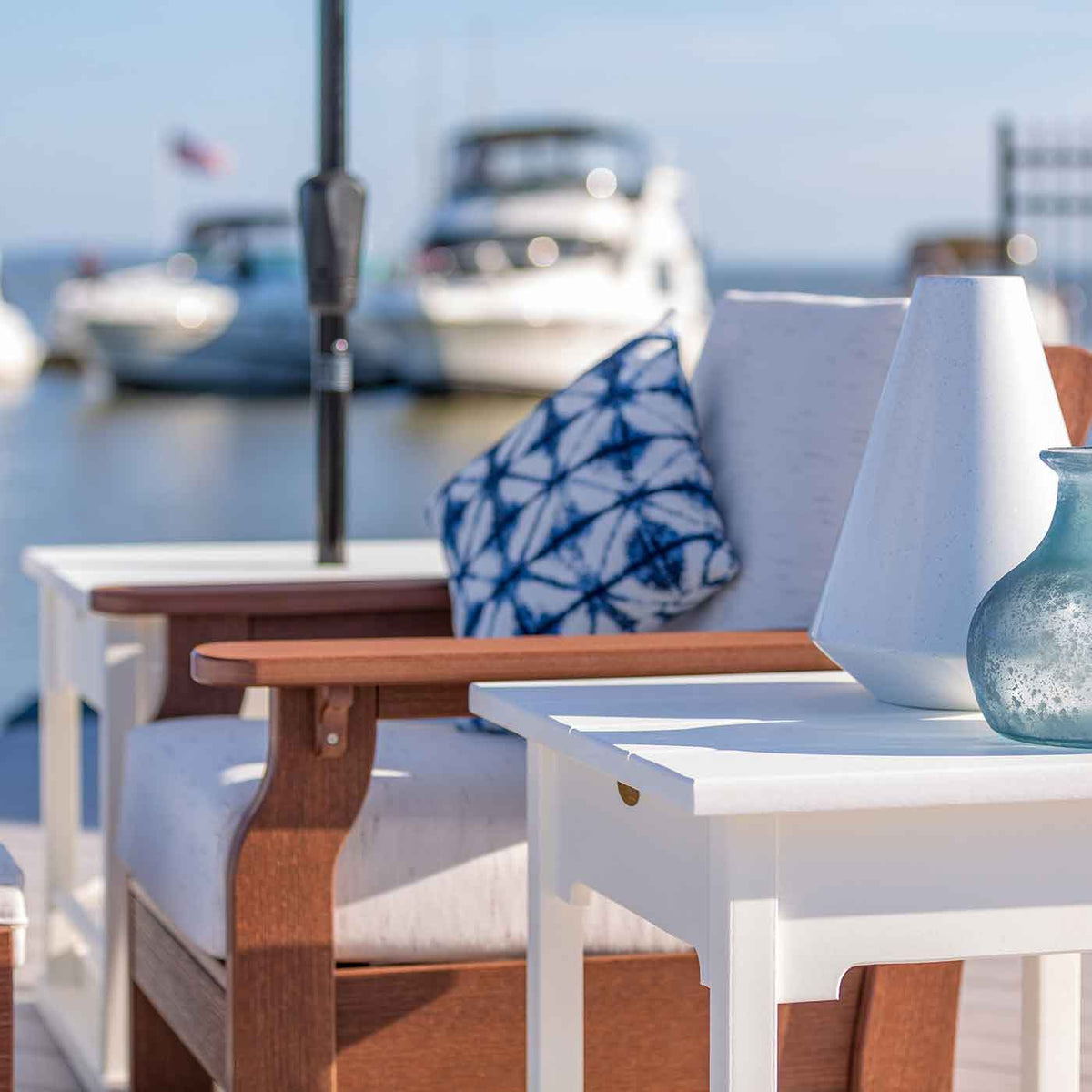 Navy tie dye outdoor pillow on a cushioned poly deck chair beside a white side table with vases at a boat marina