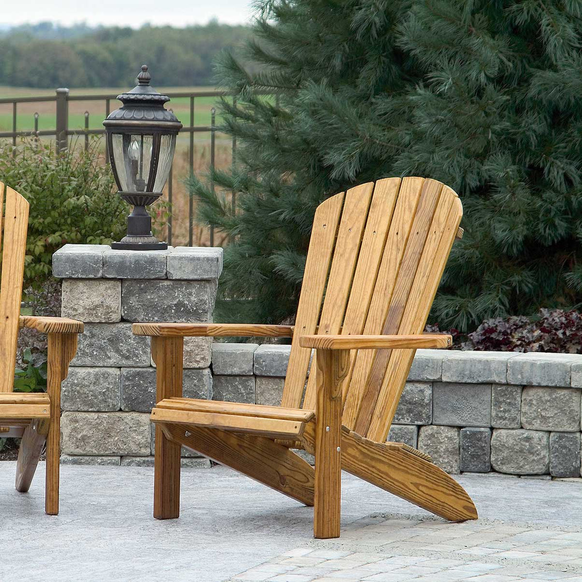 Two stained pine Adirondack chairs with fanned back slats and wide arms on a paver patio by a stone wall