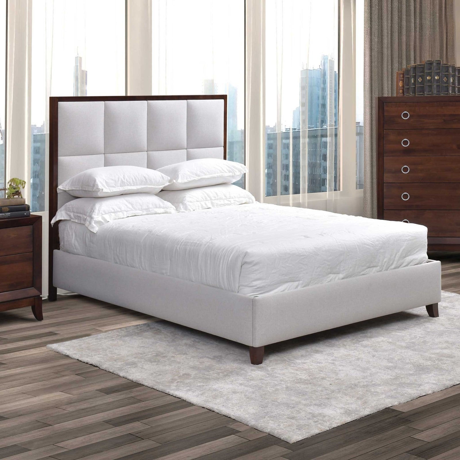 White upholstered square tufted bed with wood trimmed headboard beside a nightstand and chest in a city bedroom
