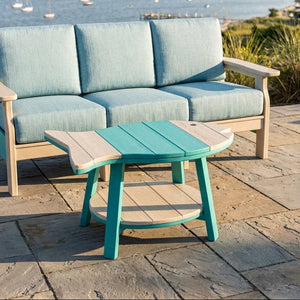 Outdoor patio furniture set with a three-seat cushioned sofa and a matching coffee table, shown on a stone deck overlooking a coastal bay with boats and shoreline.