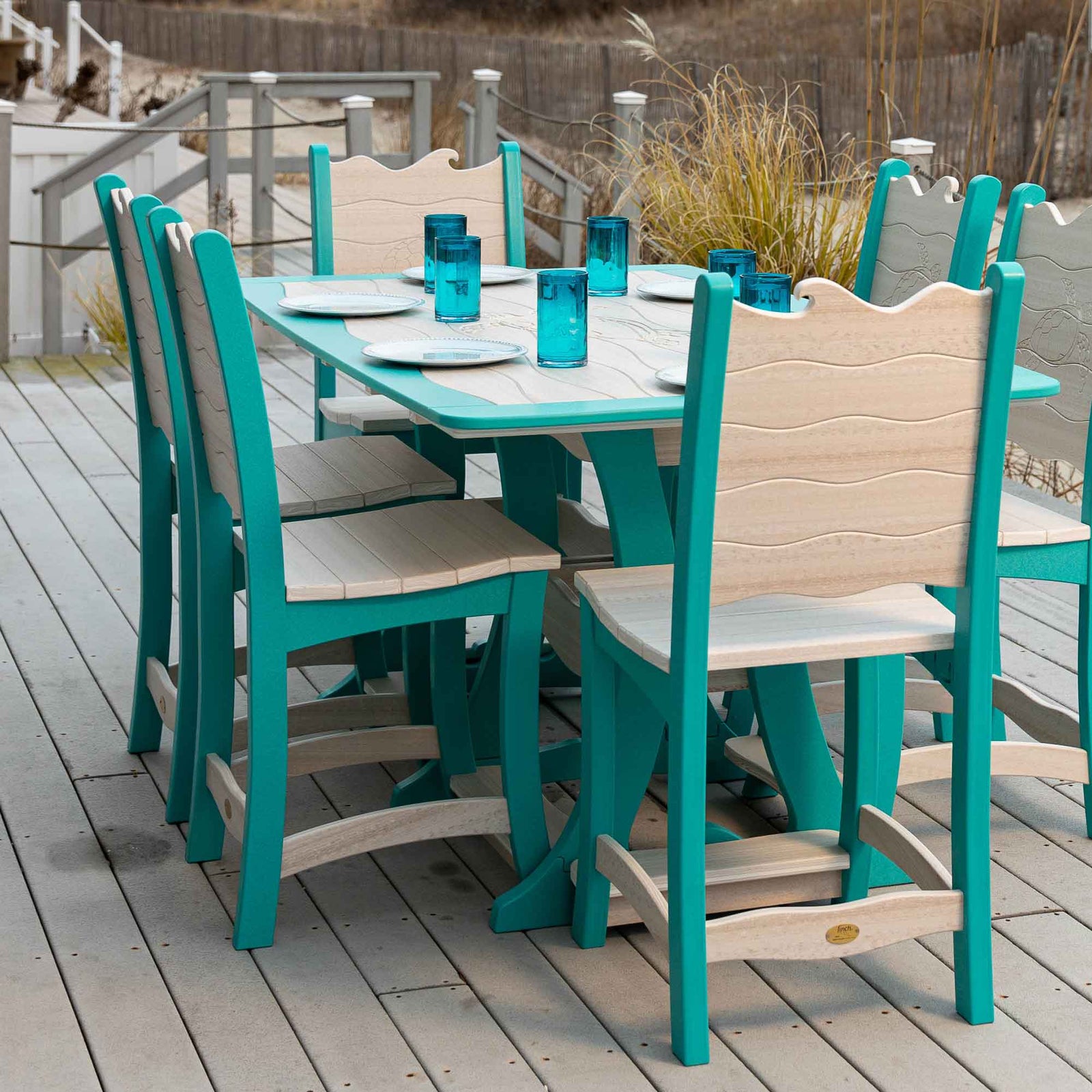 Teal and driftwood wave back counter chairs around a poly dining table set with blue tumblers on a beachfront deck