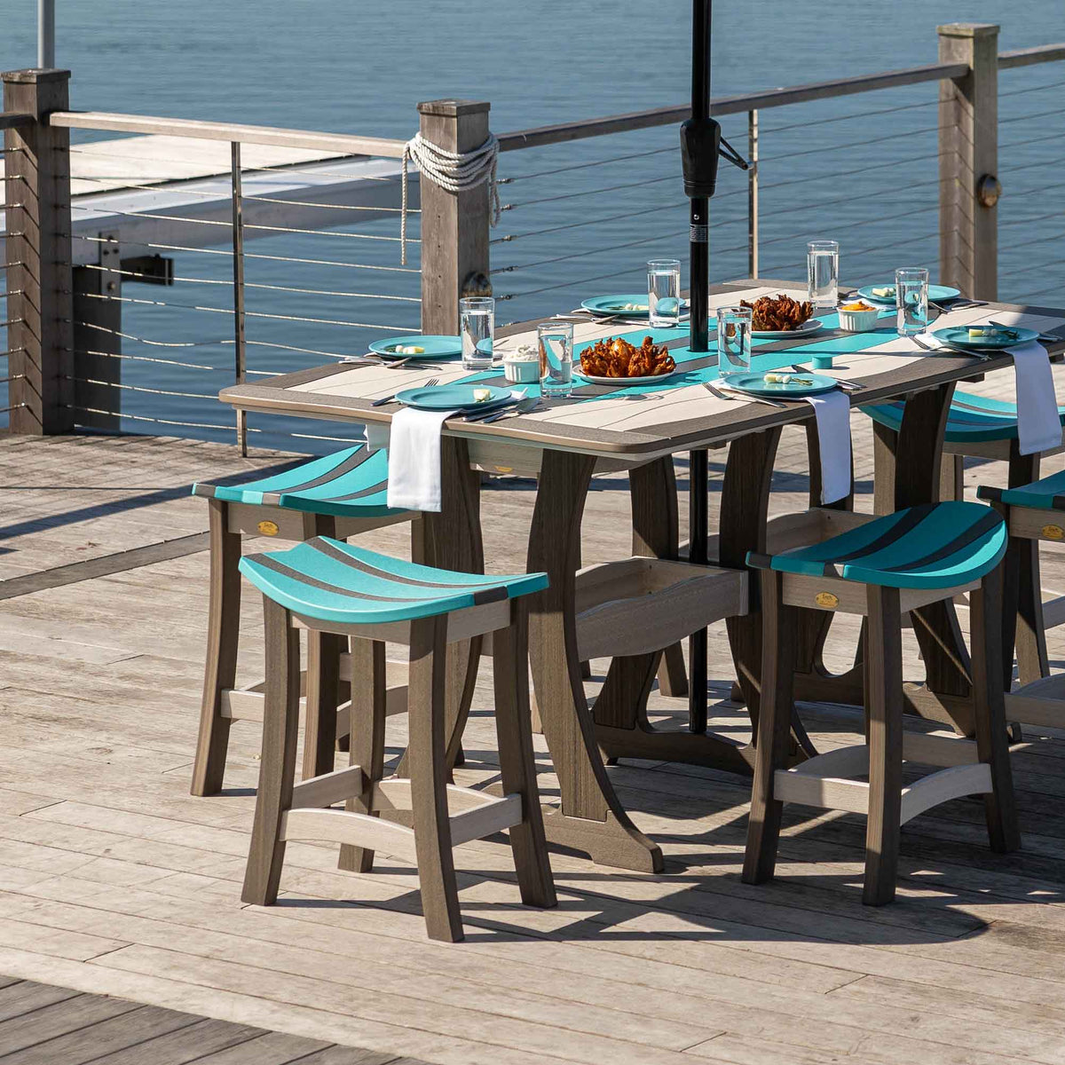 Weatherwood poly bar table with aruba blue saddle stools set with plates and food on a dock beside the water