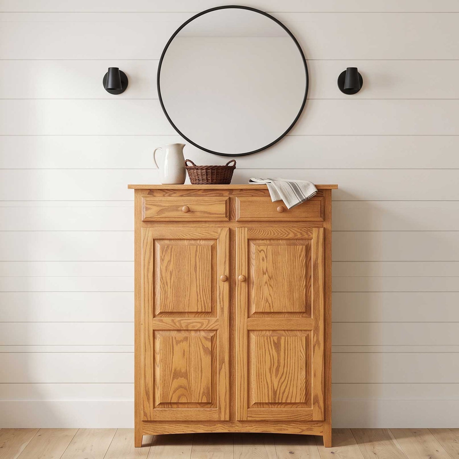 A light oak wood storage cabinet with two doors and a top drawer, styled with a white pitcher, a small basket, and a folded striped cloth on its surface, set against a white shiplap wall with a round black-framed mirror and two black wall sconces.