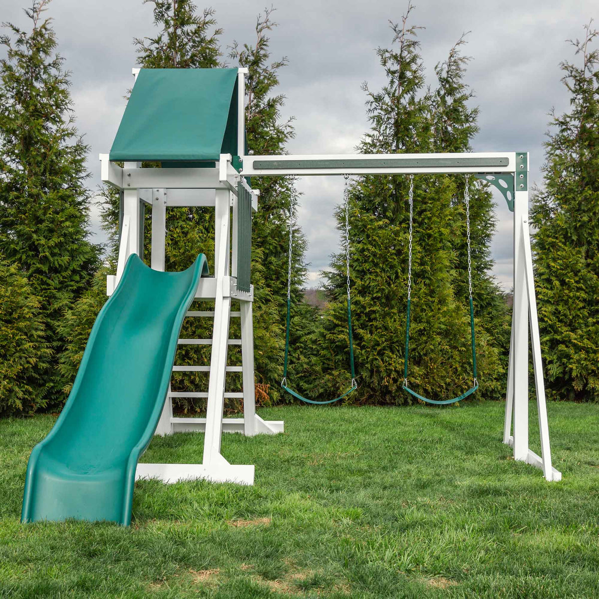 White vinyl swing set with green tarp roof, wave slide and two belt swings in a yard lined with arborvitae