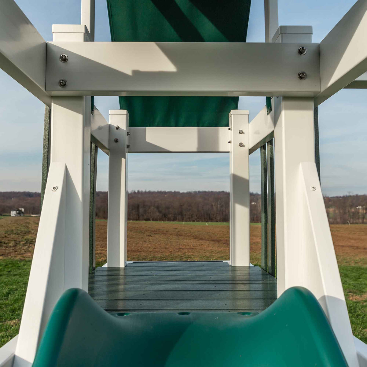 View from the playset deck through the shaded fort toward a farm field, with the green wave slide in front